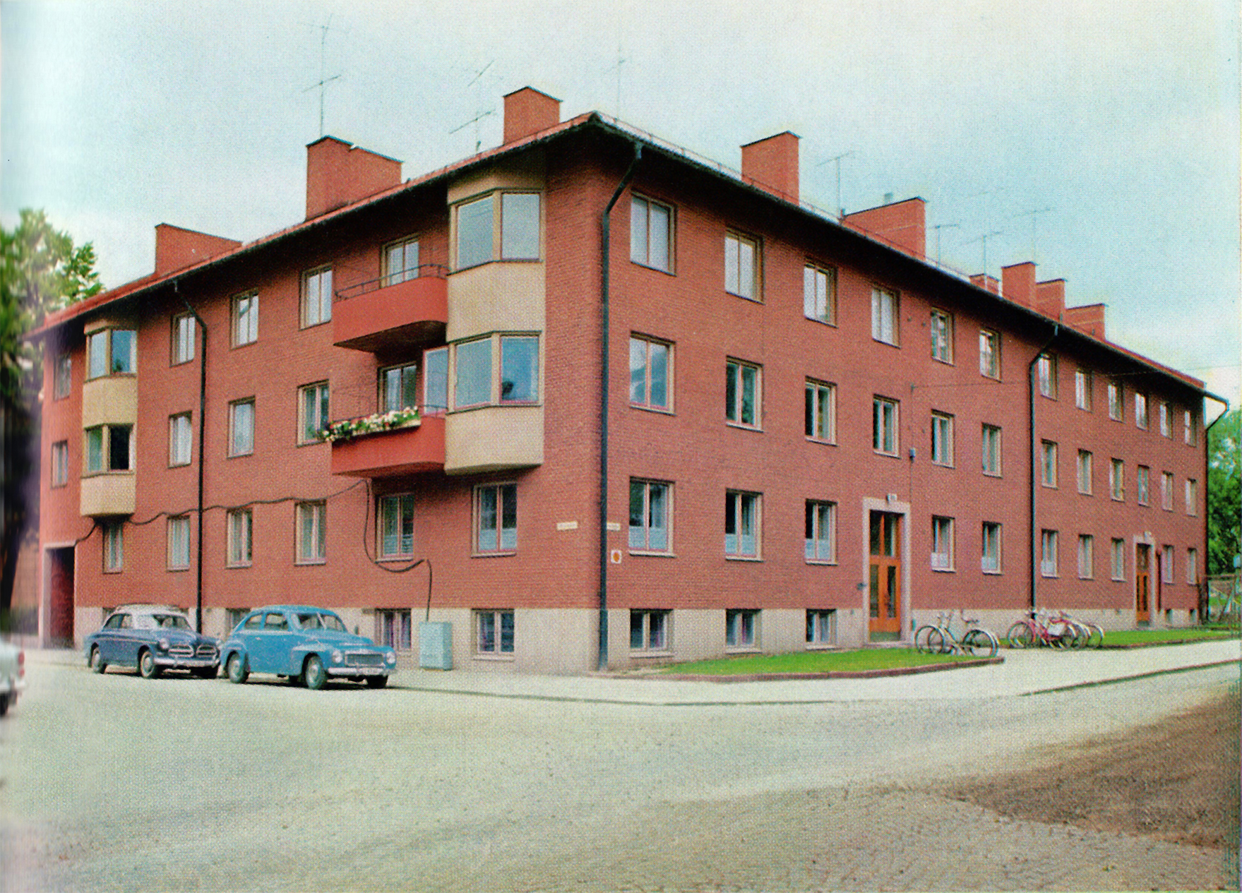 Bild på gaveln av ett tegelfärgat trevåningshus med tre portar och två balkonger. På gatan framför huset står två blå bilar parkerade.